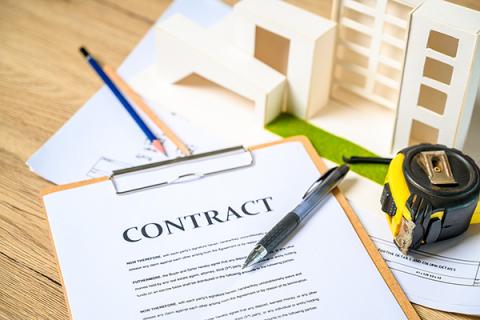 A construction contract on a table next to a tape measure and small-scale wooden buildings