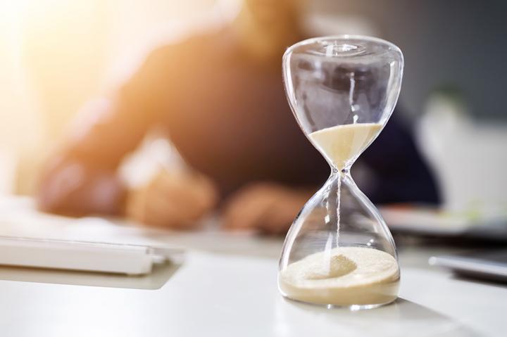 An hourglass, which is nearly out of sand, in front of a man with a stack of invoices