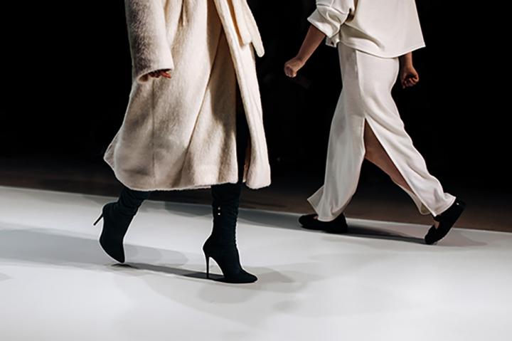 Close up of the legs of two models walking on a runway. The foreground model is wearing a white coat and black heeled boots, and the background black loafers and a white trouser suit.