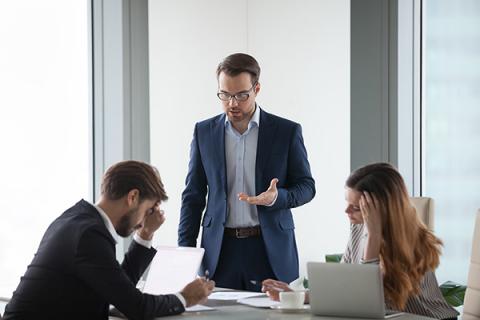 An office scenario with one man standing and a man and a woman sitting, all looking stressed