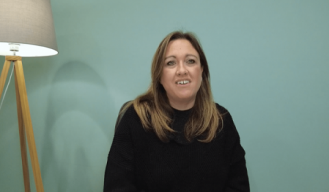 Alison Walton looking at the camera, sitting in front of a green wall with a lamp in the corner