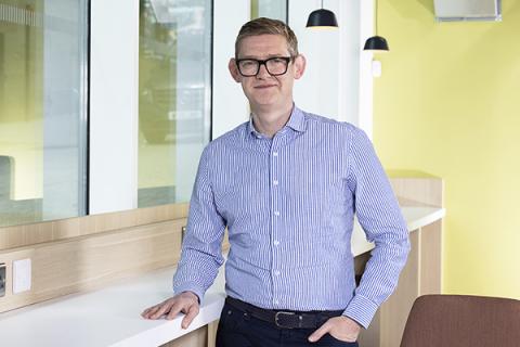 Anthony Evans wearing a blue striped shirt, leaning on a desk and smiling at the camera