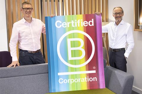 Anthony Evans and Hugh Welch, both in white shirts and dark trousers, standing behind a sofa holding a rainbow B Corp sign