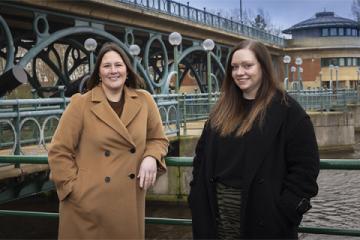 Gail Bennett, wearing a light brown coat, and Sam Alderton, wearing a black coat, standing in front of the River Tees